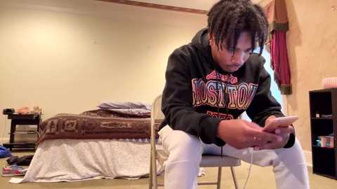 Media: Video of a young Black man with dreadlocks wearing a black hoodie and white pants, intently playing a video game on a smartphone, sitting in a cluttered, beige room with a bed and bookshelf.