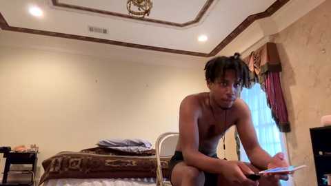 Media: Video of a shirtless Black man with dreadlocks, wearing shorts, sitting on a bed in a dimly lit room, holding a tablet.