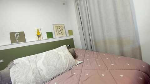 Media: Video of a minimalist bedroom featuring a green headboard, light gray curtains, a white pillow, and a beige bedspread with small heart patterns.