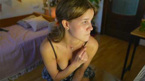 Media: Video of a young Caucasian woman with fair skin and light brown hair, wearing a black spaghetti strap top, kneeling on a wooden floor in a cozy bedroom with a bed, wooden furniture, and warm lighting.