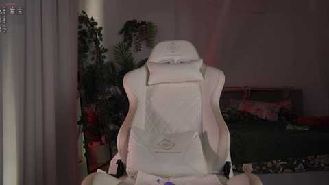 Media: Video of a white gaming chair with a cushioned headrest in a dimly lit bedroom, featuring a green bed with floral pillows, a potted plant, and a partly visible window.