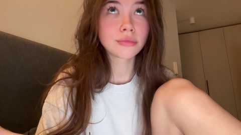 Media: Video of a young woman with long brown hair, wearing a white shirt, seated indoors. She has light skin, blue eyes, and a neutral expression. Background includes a dark couch and white wardrobe.