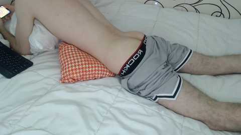 Media: Video of a fair-skinned, shirtless man lying on his stomach on a bed, wearing gray athletic shorts with a black waistband. He's holding a smartphone. The bed has a white quilt and an orange pillow with a houndstooth pattern.