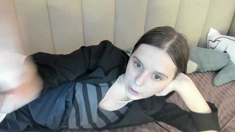 Media: A video shows a young woman with light skin and dark hair lying on a bed, wearing a black and grey striped top, with a cushion beside her.