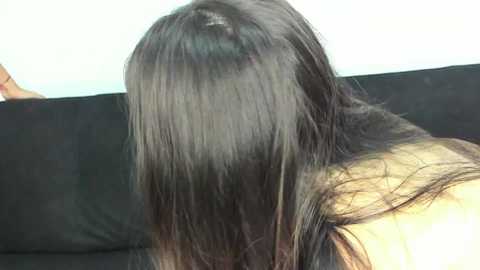 Media: Video of a person with long, straight black hair covering their face, sitting on a black couch against a light wall.