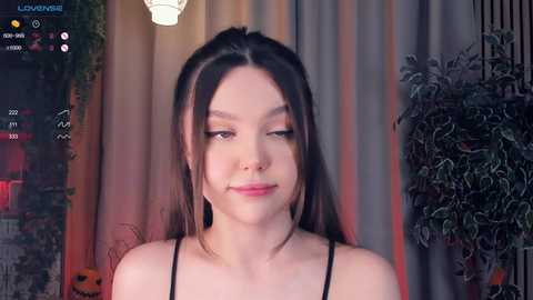 Media: A video of a young woman with long dark hair, fair skin, and closed eyes, wearing a black top, standing in front of a Halloween-themed background with a potted plant, a pumpkin, and a curtain.