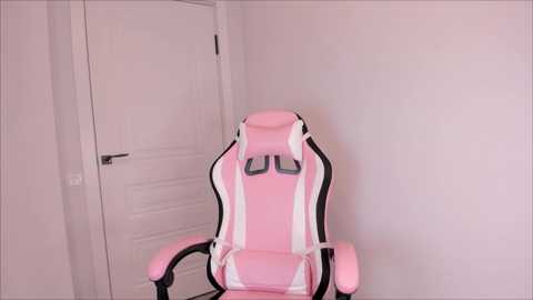 Media: Video of a pink and white gaming chair with adjustable armrests, situated in a minimalist room with white walls and a closed door.