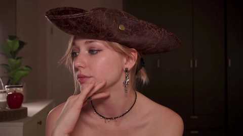 Media: Video of a blonde woman in a dark hat, wearing a black necklace, sitting in a kitchen with a red apple on a counter.