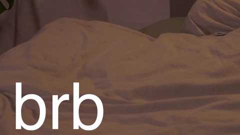 Media: A video of a person's upper body, wrapped in a white blanket, lying on a bed with a pillow. The text \"brb\" is superimposed in white on the left side. The background is dimly lit, creating a cozy, intimate atmosphere.