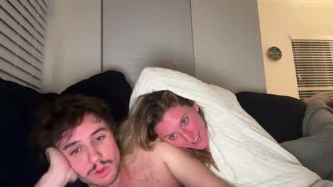 Media: Video of a man and a woman lying on a dark couch under a white blanket. The woman, with blonde hair, is partially visible, smiling. Background shows a beige wall with a window and blinds.