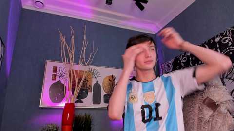 Media: Video of a young man in a white and blue soccer jersey, hands covering face, in a dimly lit room with blue walls, abstract art, and a red vase.