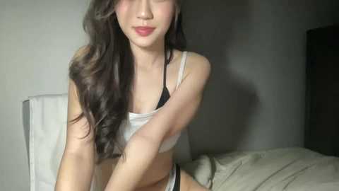 Media: Video of a slender, fair-skinned Asian woman with long, wavy dark hair, wearing a white tank top and black thong, sitting on a bed with beige sheets, in a dimly lit, simple bedroom.