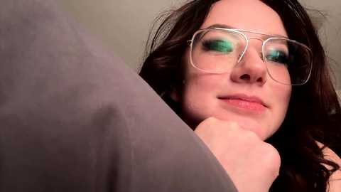 Media: Video of a woman with fair skin, dark brown hair, and green eyeshadow, wearing large, round glasses and resting her chin on her hand, with a grey sleeve in the foreground.