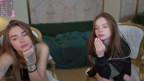 Media: Video of two young women with long brown hair, one with a tattooed arm, sitting on a green couch, wearing black clothing, in a luxurious room with gold accents.