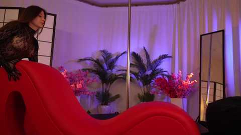 Media: Video of a woman in a black lace top, leaning on a red leather chaise lounge, surrounded by potted pink flowers and tall green plants, in a dimly lit, modern room with white curtains.