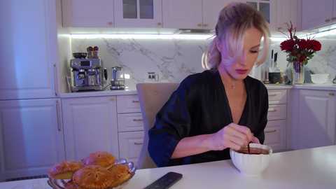 Media: Video of a blonde woman in a black robe, seated at a white kitchen table, eating soup from a white bowl, surrounded by pastries and a vase of red roses.