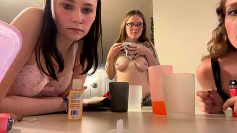 Media: Video of three young women with fair skin and medium-sized breasts, one topless, sitting around a table with various objects, including a phone, in a dimly lit room.