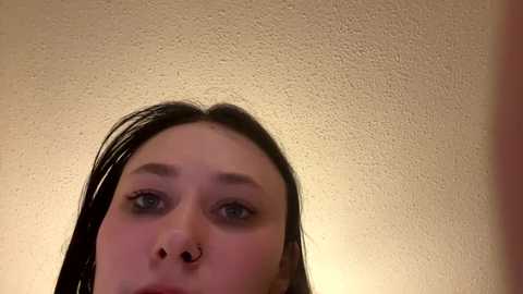 Media: Video of a young woman with long, straight dark hair, pale skin, and blue eyes, looking up. She has a septum piercing and wears minimal makeup. The background is a textured beige wall.