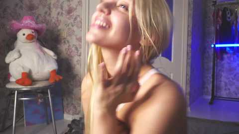 Media: Video of a smiling, nude blonde woman with small breasts, touching her neck, in a room with floral wallpaper, a stuffed duck, and a blue light.