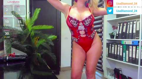 Media: Video of a blonde woman in a red lace bodysuit, posing confidently in a modern office with a calendar, green plant, and bookshelves in the background.