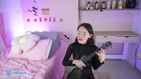 Media: Video of a young Asian woman with long black hair and glasses, wearing a black top, playing an electric guitar in a pink-themed bedroom with a white bed, stuffed animals, and wall decorations.