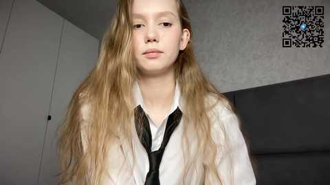 Media: Video of a young, fair-skinned girl with long, wavy blonde hair, wearing a white shirt and black tie, indoors against a gray wall.