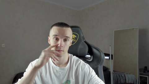 Media: A video of a young man with short brown hair, wearing a white T-shirt, seated in a black gaming chair with yellow accents, in a sparsely furnished room with beige walls and a mirror.