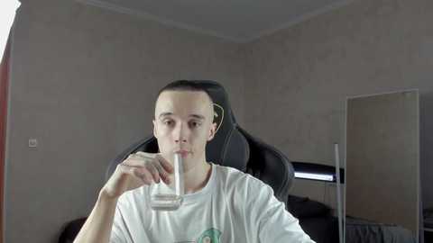 Media: Video of a young man with short dark hair, wearing a white T-shirt, drinking from a glass, seated in a black gaming chair in a simple, gray-walled room.