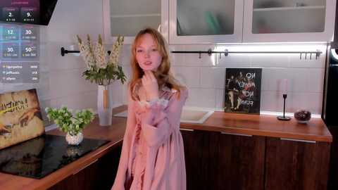 Media: Video of a young, fair-skinned woman with long, wavy blonde hair, wearing a light pink, off-the-shoulder dress, standing in a modern kitchen with wooden cabinets, glass-front doors, and a black countertop.