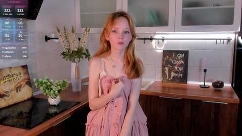 Media: Video of a young, fair-skinned, red-haired woman in a pink dress standing in a modern kitchen with wooden cabinets, glass doors, and a white subway tile backsplash.