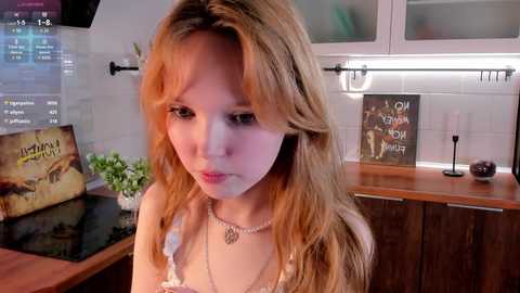 Media: Video of a young woman with fair skin and long, wavy blonde hair in a kitchen, wearing a white lace bra and a pearl necklace.