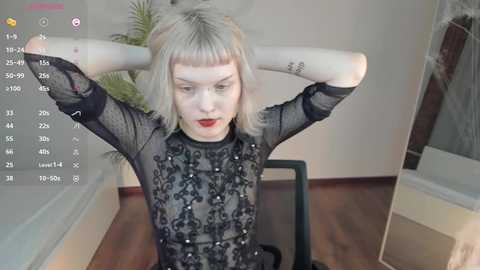 Media: A video of a pale-skinned, blonde-haired woman with a bob haircut, wearing a sheer black lace dress, sitting on a chair, arms behind her head, in a modern room with a window and a plant.