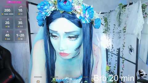 Media: A video of a pale-skinned woman with blue hair, wearing a blue flower crown and a white veil, set against a white background with spiderweb decorations, overlayed with a video recording interface.