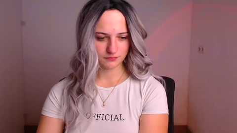 Media: A video of a young woman with long, wavy gray hair, wearing a light gray \"Official\" T-shirt, seated indoors with a blurred background.