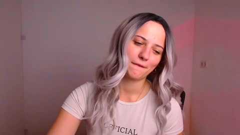 Media: Video of a fair-skinned woman with long, wavy, silver-grey hair, wearing a white t-shirt, smiling gently. Background is a plain, light-colored wall with soft pinkish lighting.