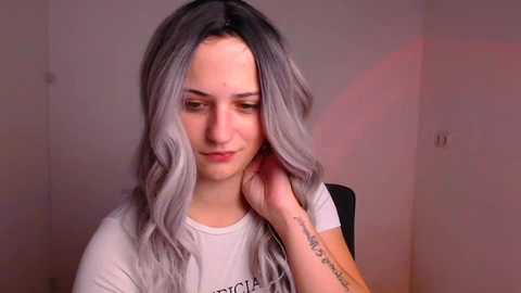 Media: Video of a young woman with wavy, silver-gray hair, fair skin, wearing a white t-shirt. She's seated, looking down, with a contemplative expression. Background is a plain, light-colored wall.