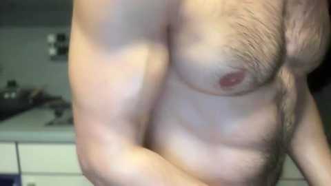 Media: Video of a light-skinned man with a hairy chest and medium build, standing shirtless in a dimly lit bedroom. The background shows a bed with indistinct objects.