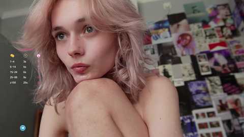 Media: A video of a young, fair-skinned woman with shoulder-length, wavy, pastel pink hair, wearing nothing but a sultry expression. Background shows a cluttered wall of photos and posters.