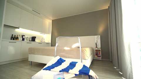 Media: Video of a modern, minimalist bedroom with a white bed, beige headboard, and light wooden floor. A blue and white striped pillow lies on the floor in front. White cabinets, kitchen area, and a small bookshelf are visible.