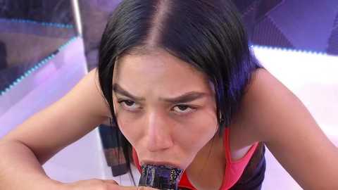 Media: Video of an Asian woman with long black hair, fair skin, and a slender physique, performing oral sex on a black penis, wearing a red tank top. The background is a modern, dark room with blue and purple lighting.