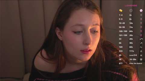 Media: Video of a young woman with fair skin and straight brown hair, wearing a black off-shoulder sweater, looking introspective against a beige background.