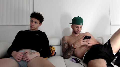 Media: Video of two shirtless men, one with a green cap, sitting on a white couch, one wearing black shorts, one with tattoos, both looking at smartphones.