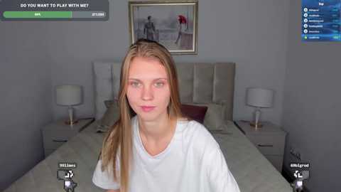 Media: A video of a young, fair-skinned, blonde woman with light makeup, wearing a white T-shirt, sitting on a bed in a modern, minimalist bedroom. The room has gray walls, a framed painting, and white lamps on nightstands.