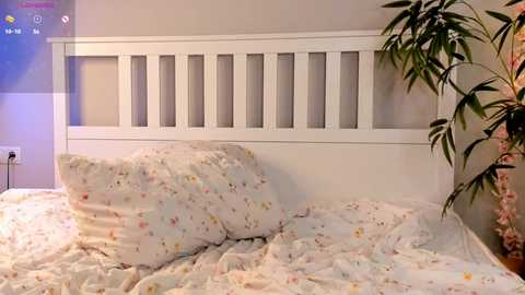 Media: Video of a cozy bedroom with a white wooden headboard, floral-patterned bed linens, a tall potted plant, and soft, ambient lighting.