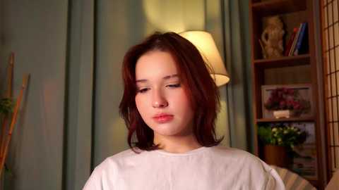 Media: Video of a young Asian woman with shoulder-length auburn hair, wearing a white shirt, indoors near a green curtain, a wooden shelf with books and plants, and a lit lamp.