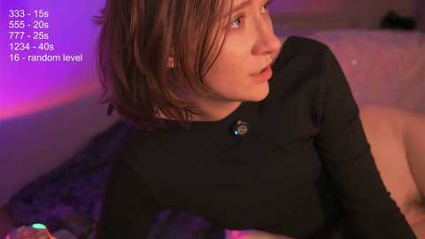 Media: A video of a young, fair-skinned woman with shoulder-length brown hair, wearing a black long-sleeved top, seated on a bed with purple lighting, revealing a 353/5-265/25 random level.