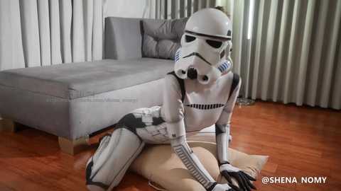 Media: Video of a person in a white, detailed Star Wars Stormtrooper costume, sitting on a beige cushion on a wooden floor, with a grey sectional sofa in the background, and light grey curtains.