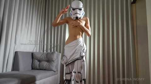 Media: Video of a shirtless man wearing a Star Wars Stormtrooper helmet, draped in a white towel, standing in a modern living room with grey curtains and a grey sofa.