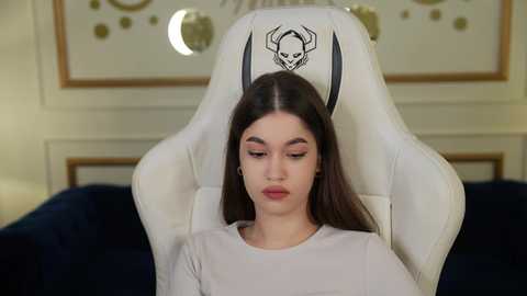 Media: Video of a young woman with long dark hair, wearing a white T-shirt, reclining in a white gaming chair with a skull and horns logo. Background features a dark blue couch and a wall with circular and moon-themed art.