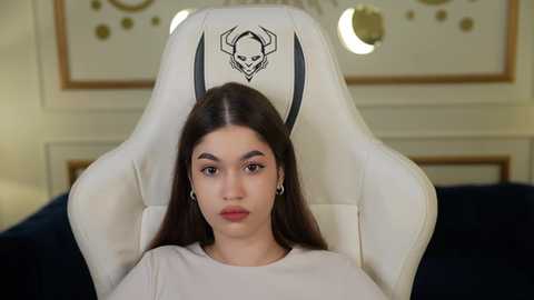 Media: Video of a young woman with long dark hair, wearing a white gaming chair with a skull and crossbones design, in a room with beige walls and round light fixtures.